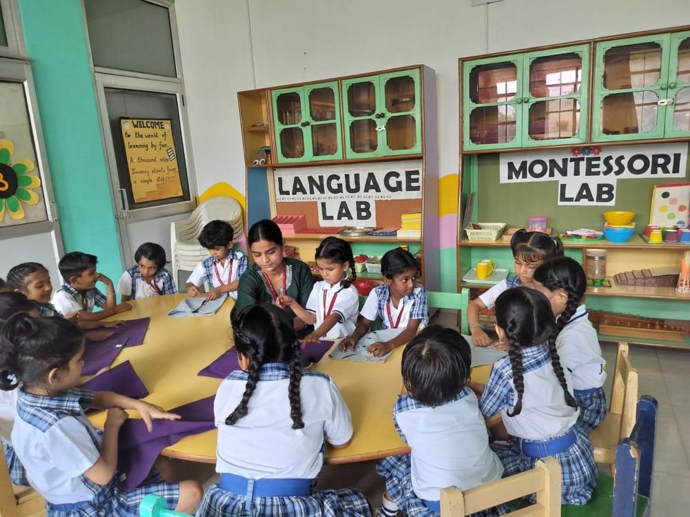 Joyland Montessori School children and their teacher seated at a yellow table handling pieces of cloth, in front of the Language Lab and Montessori Lab shelves