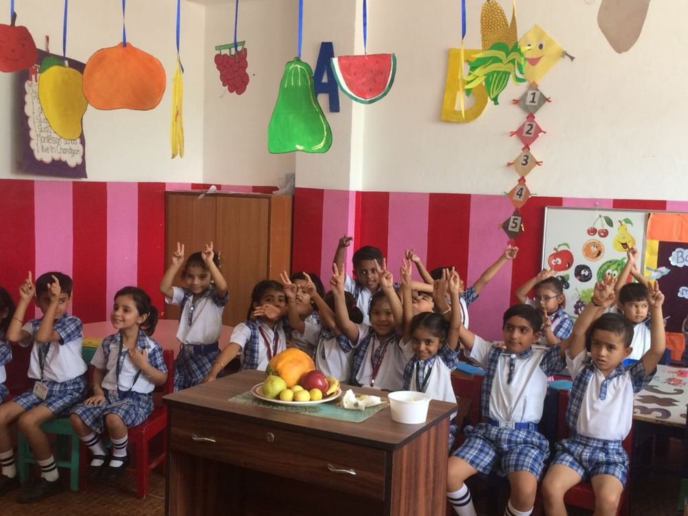 Joyland children raising their hands in a classroom beside a table of fruit